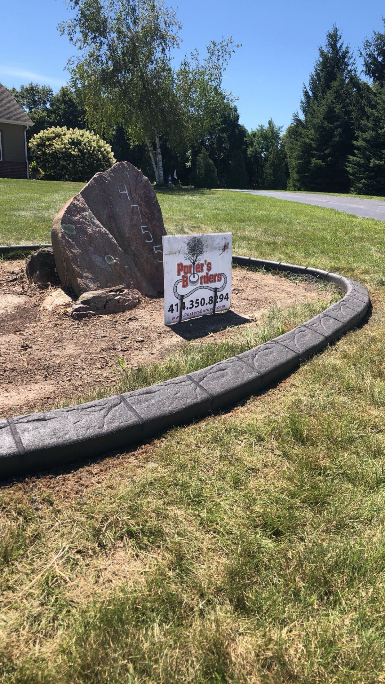 A large rock is sitting in the middle of a lush green field next to a concrete curb.