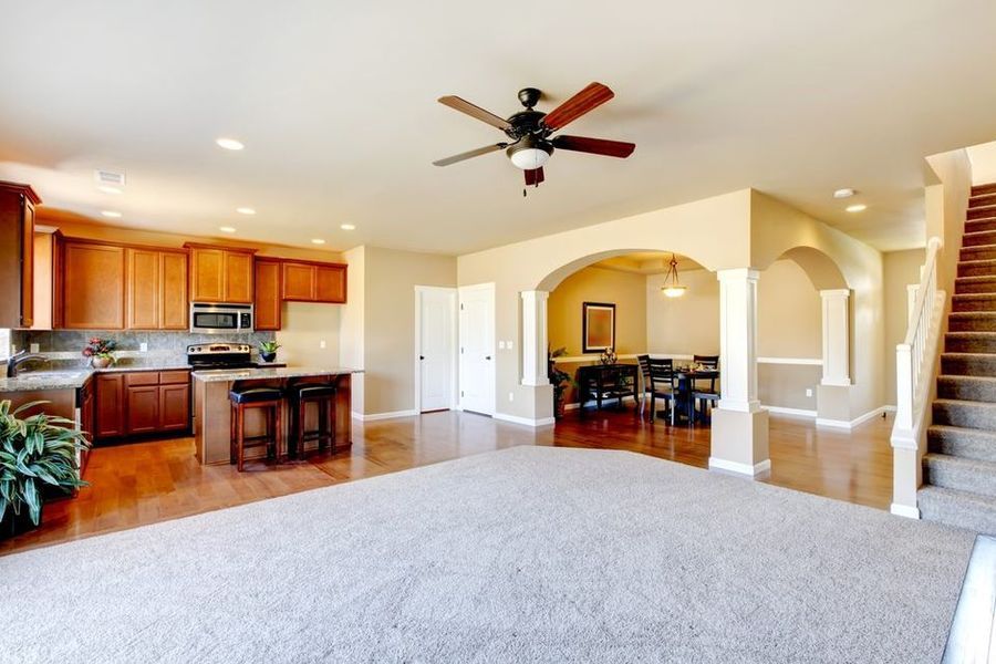 A living room with a ceiling fan and a kitchen in the background.