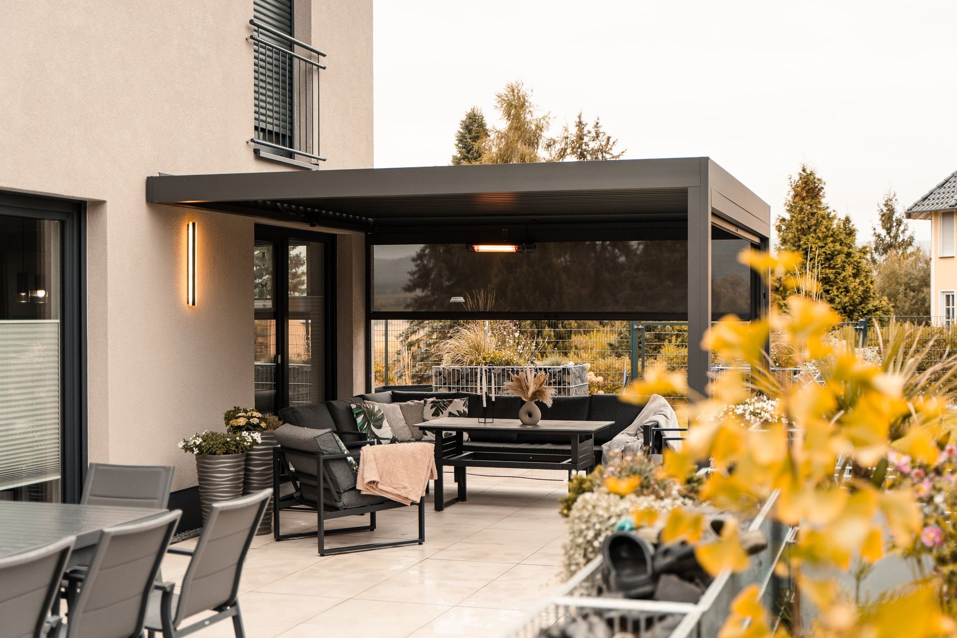 Modern patio with black pergola, outdoor seating, and warm wall lights beside a house