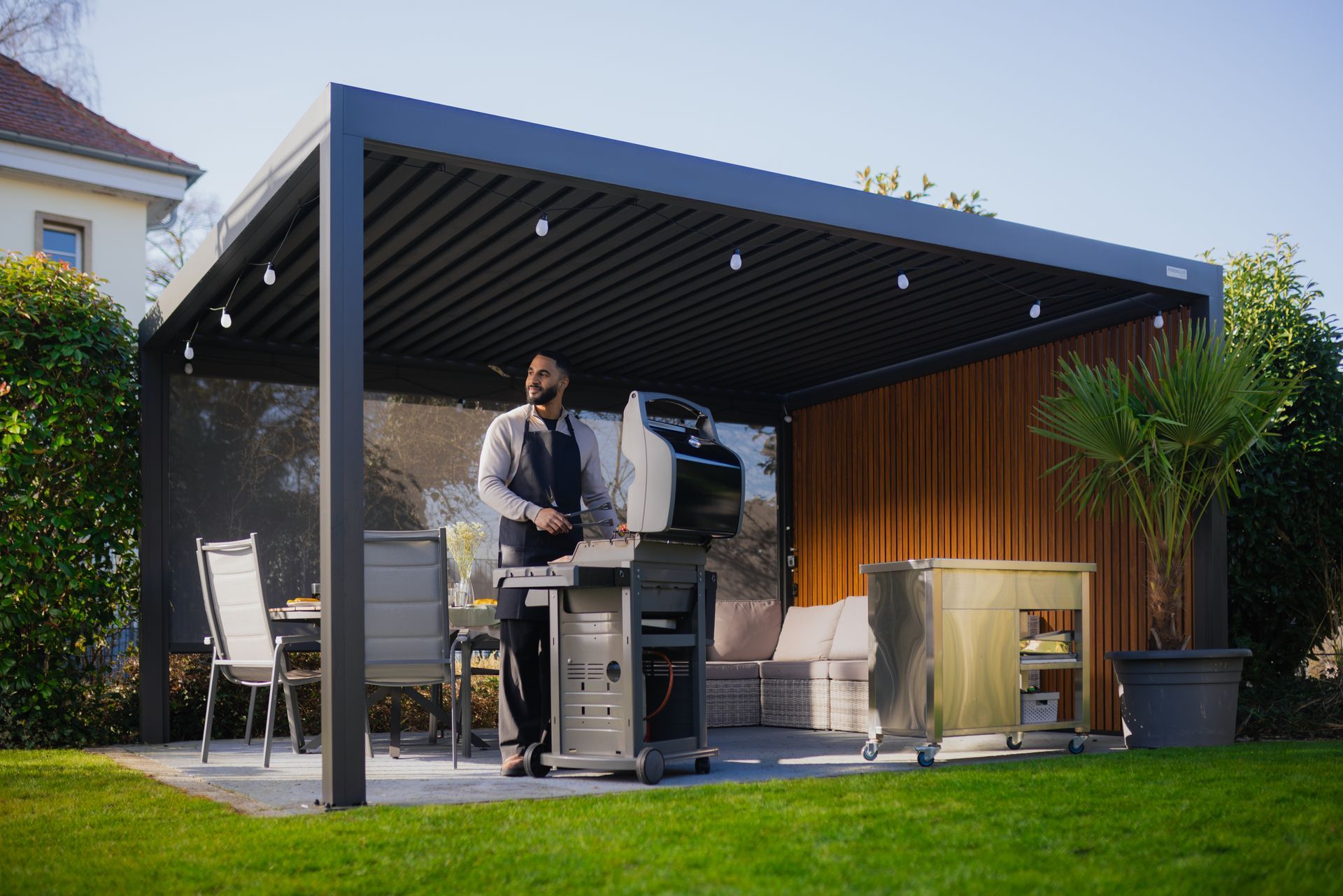 Person grilling under a modern patio roof in a backyard with lawn and potted plants