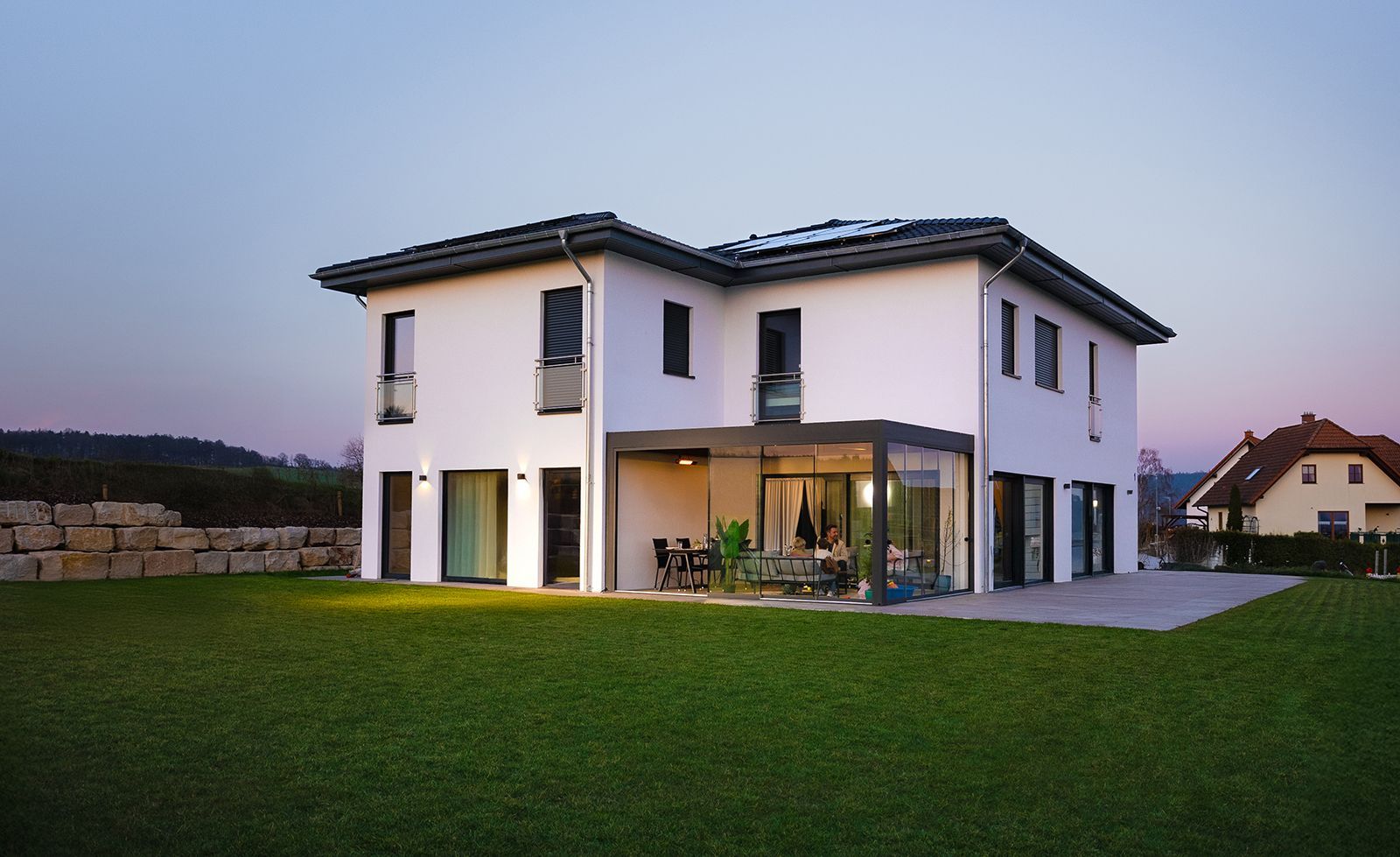 Modern white two-story house with large windows and a lit patio at dusk