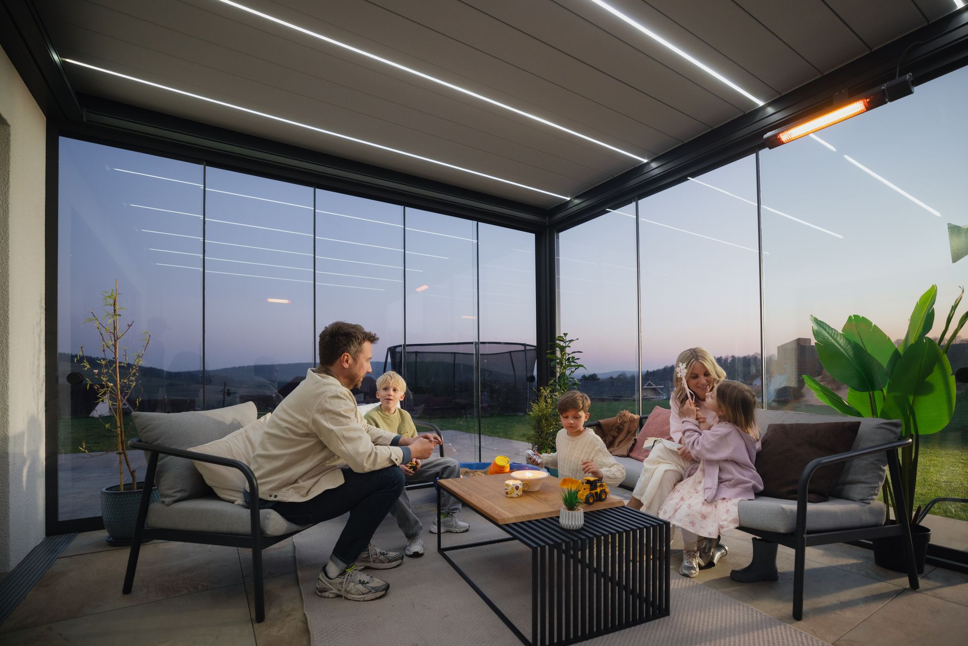 Group seated around a low table in a modern glass-walled lounge at sunset, sharing drinks and snacks.