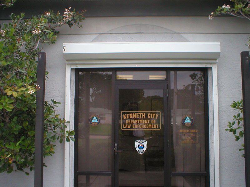 Entrance to a building with glass doors, a sign reading “Kennedy City,” and a leafy tree on the left.