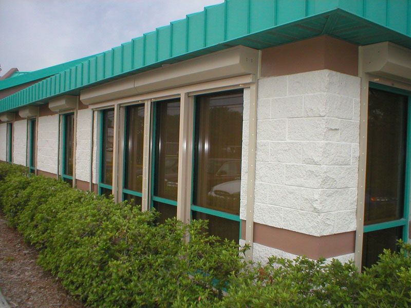 Single-story building with teal awning, large windows, and hedges along the exterior walkway