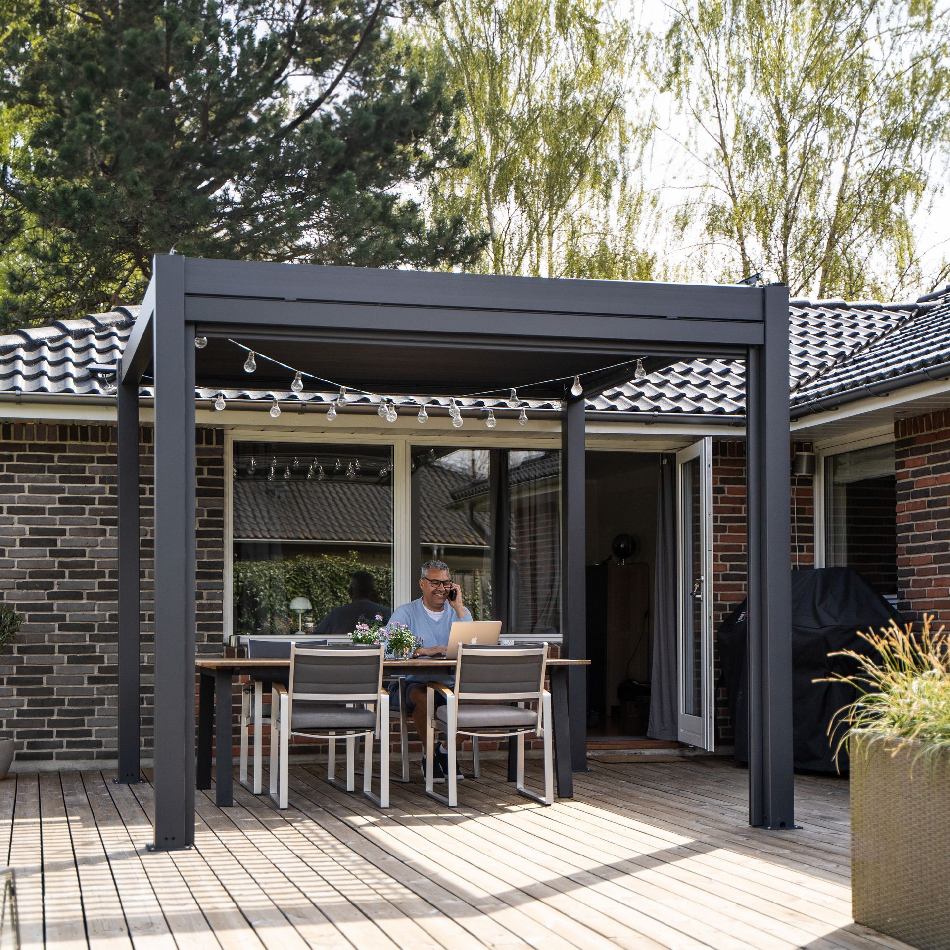 Person sitting at a patio table under a pergola in a backyard, with chairs and a brick house nearby