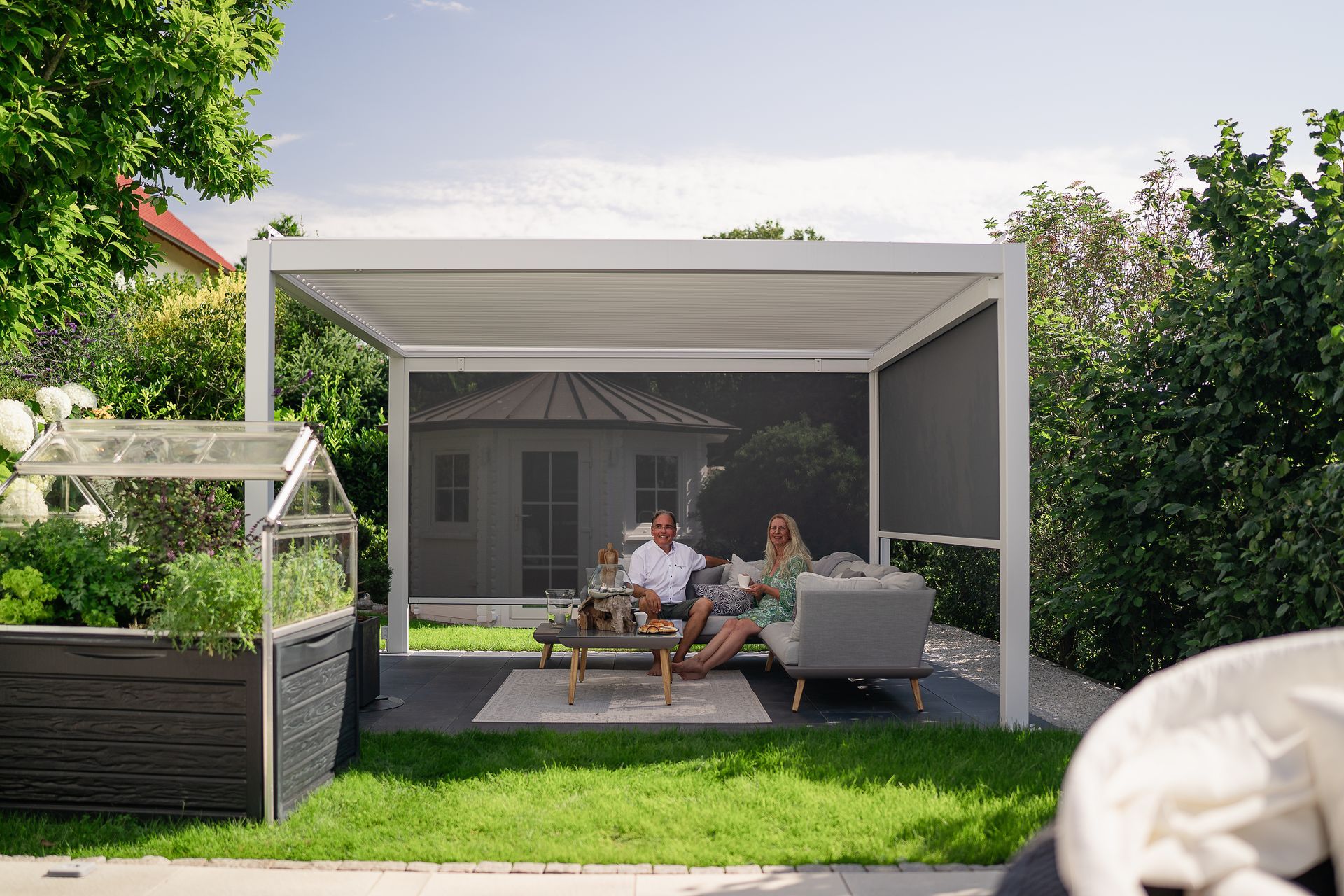 People relaxing on an outdoor patio under a white pergola beside a green lawn and garden.