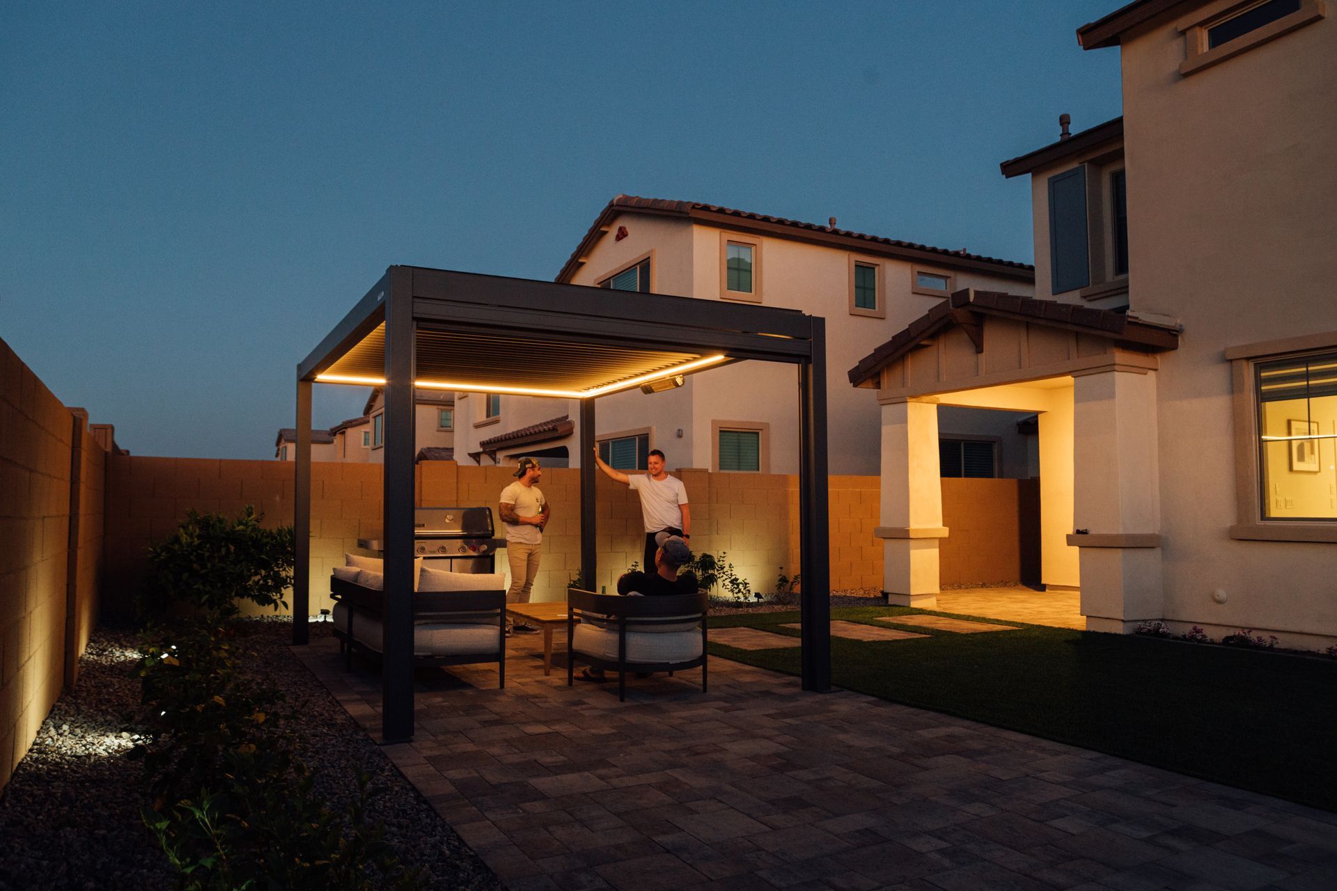 Backyard patio at dusk with a lit gazebo beside a house and two people seated inside.