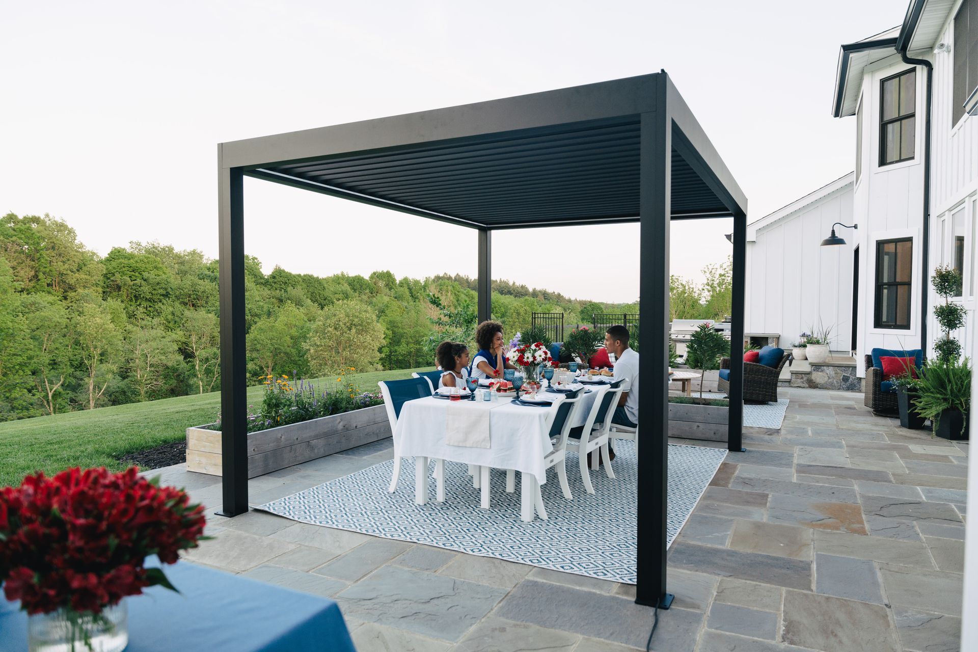 Outdoor patio dining under a black pergola beside a white house, with a table set for guests and garden views