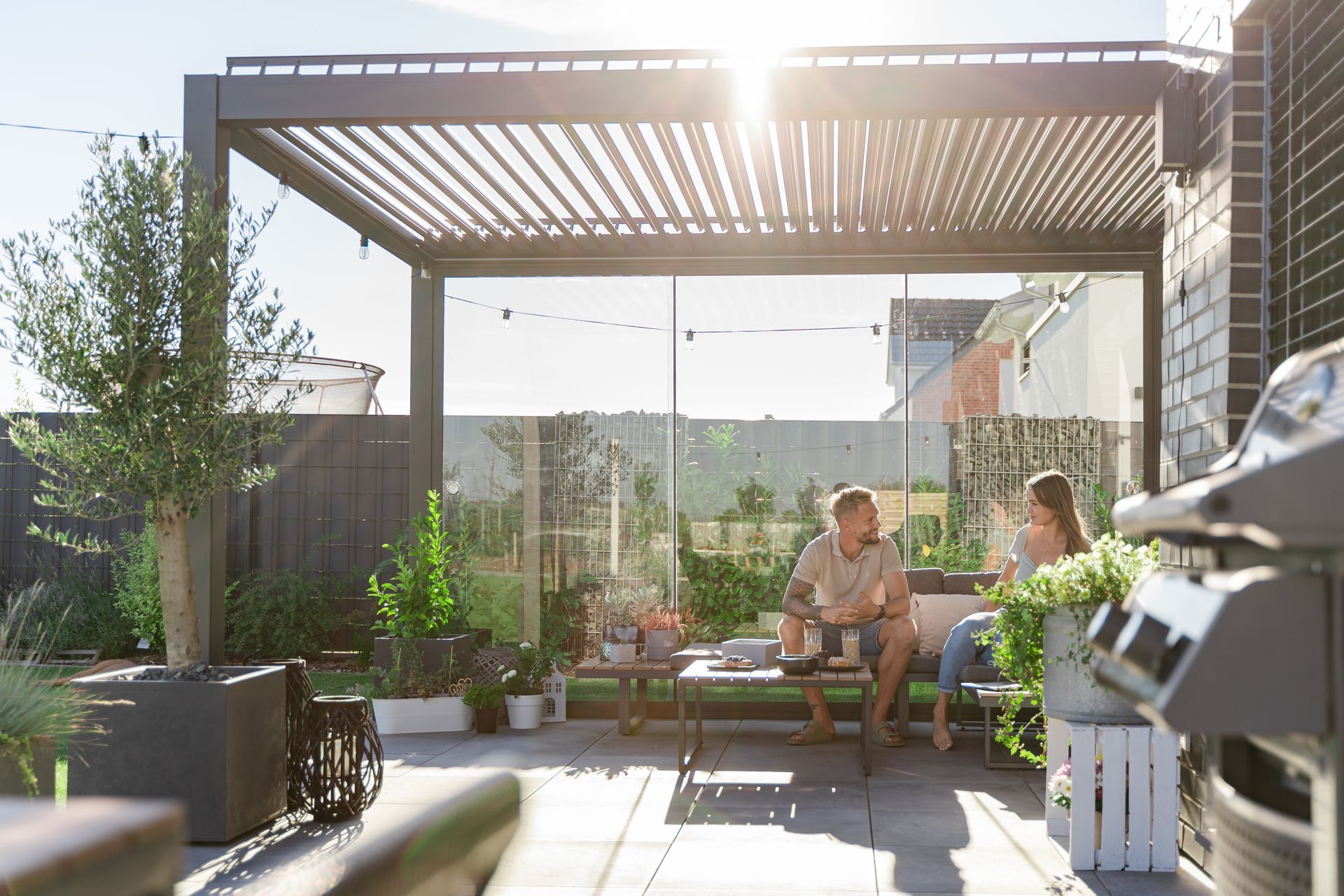 Two people sit under a slatted pergola on a sunny rooftop patio with potted plants and city views.