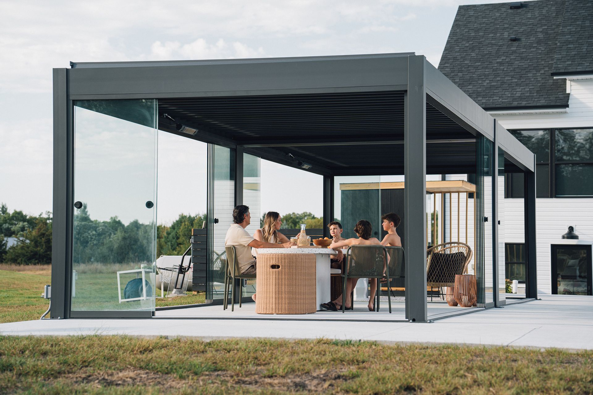 People dining under a gray patio pergola beside a modern house, with wicker chairs and a lawn beyond.