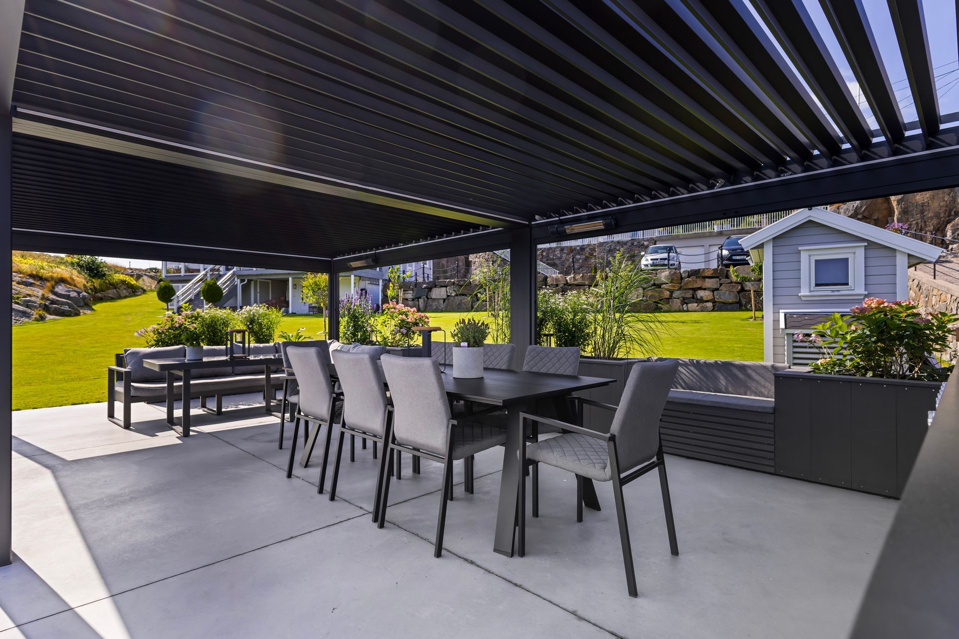Covered outdoor patio with black dining table and chairs under a slatted pergola, overlooking a landscaped yard and house.