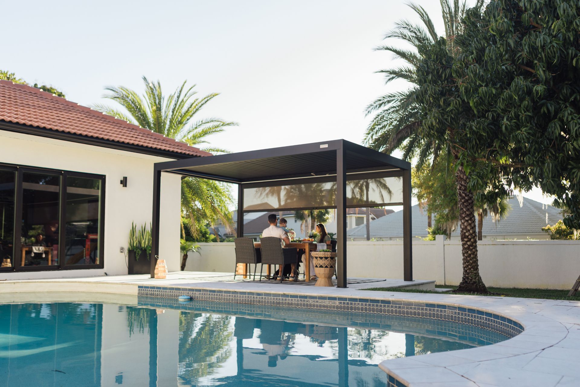 Modern backyard pool with covered patio seating and palm trees beside a white villa