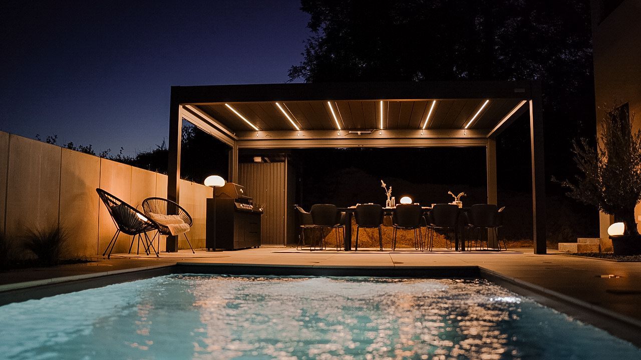 Lit backyard pool beside a modern patio lounge under warm string lights at night