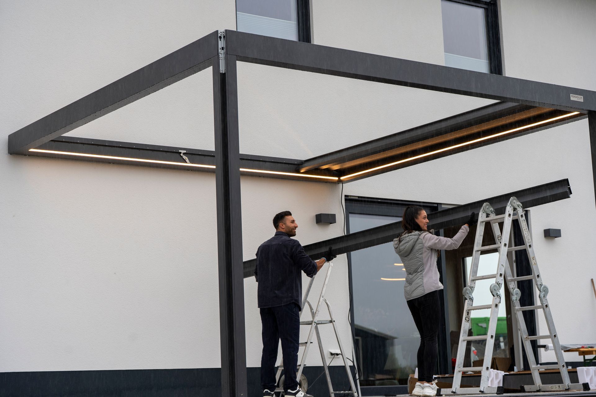Two workers assembling a large black metal pergola frame outdoors beside ladders