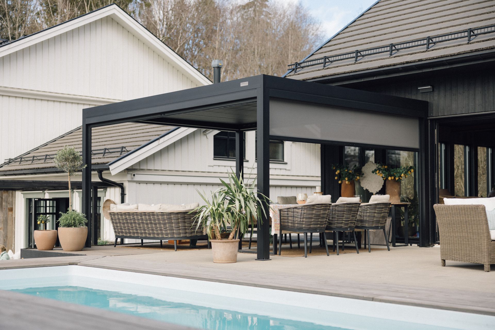 Modern backyard patio with a black pergola, dining area, potted plants, and a swimming pool.