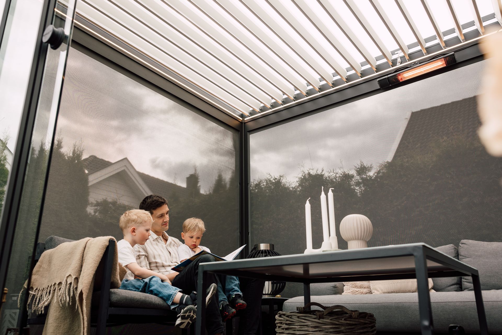 Family sitting on a patio under a pergola, with a table and candles in a modern outdoor lounge.