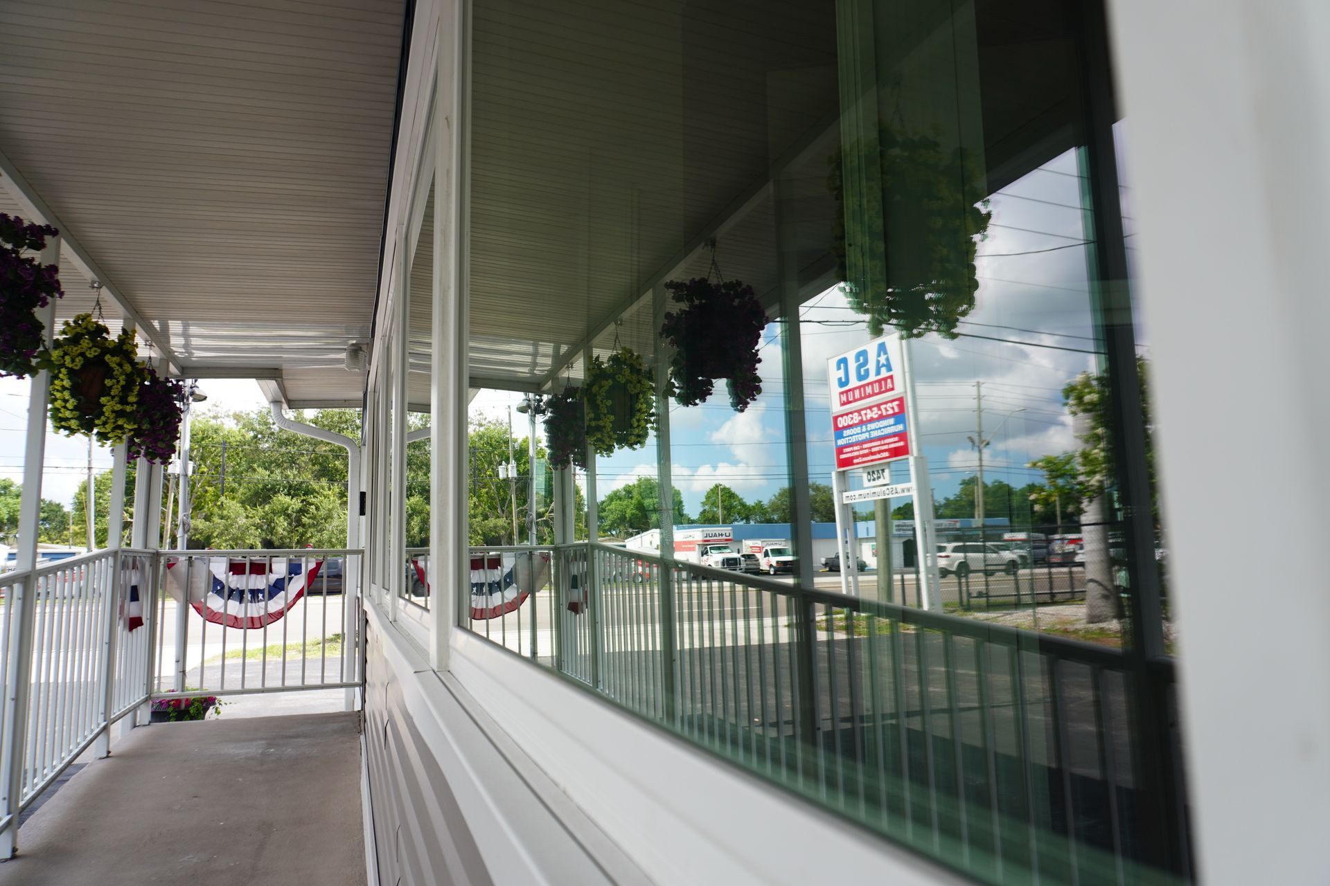 Storefront walkway with hanging flower baskets, white railings, and patriotic bunting reflected in large windows