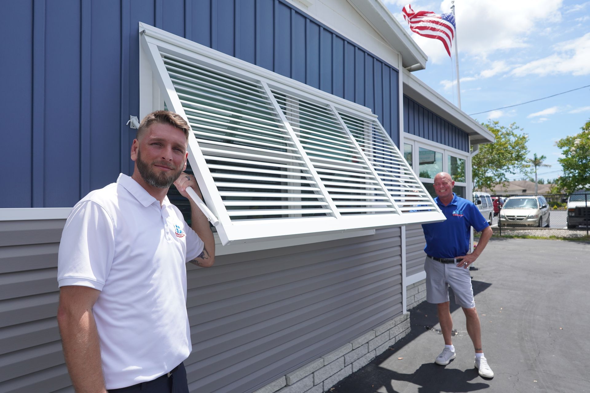 Two men standing beside a blue building and a large white awning window on a sunny street.