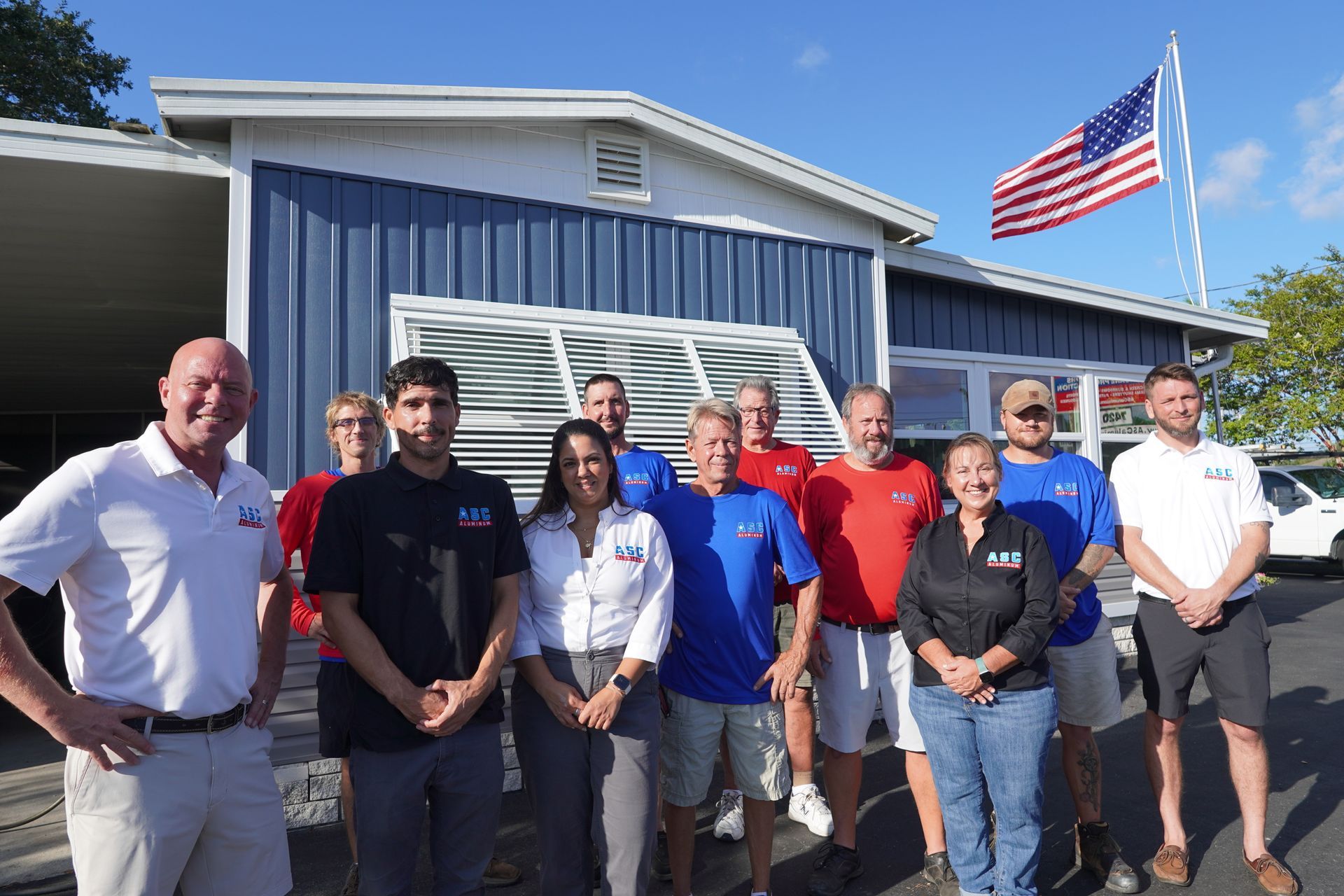 Group of people standing outside a blue building with an American flag waving overhead.