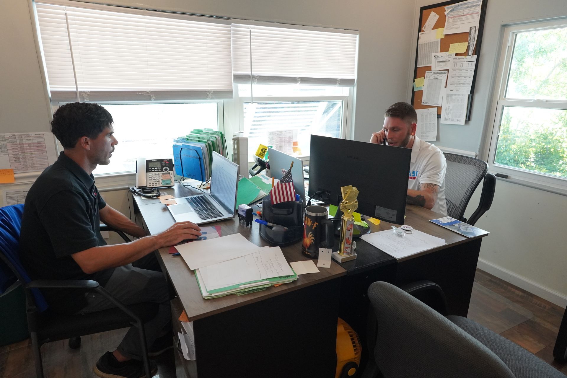 Two people working at desks in a bright office with computer monitors and papers