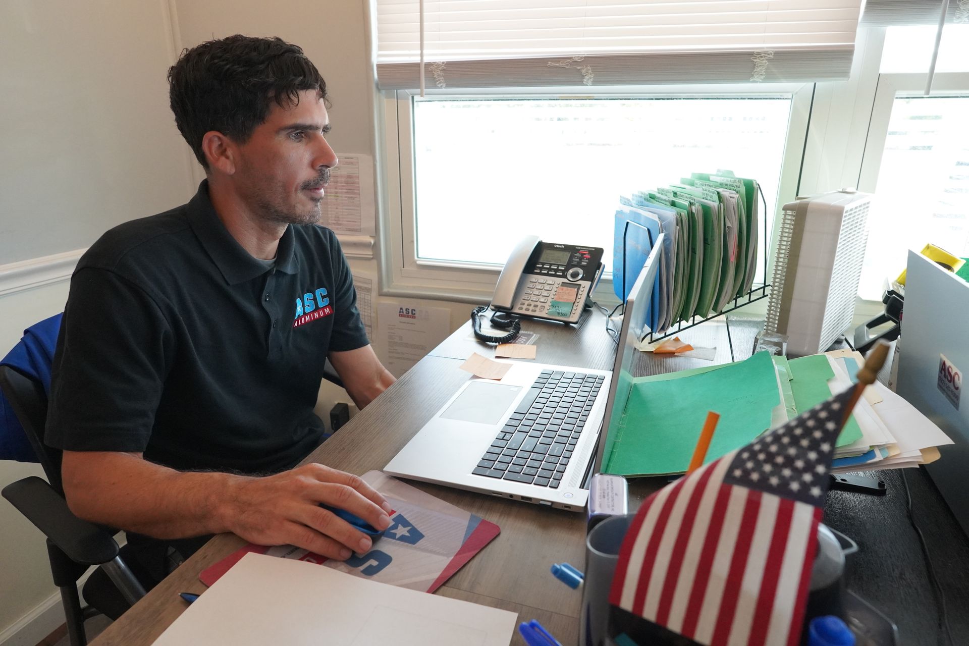 Man seated at a desk using a laptop in a bright office, with files, phone, and small US flag nearby