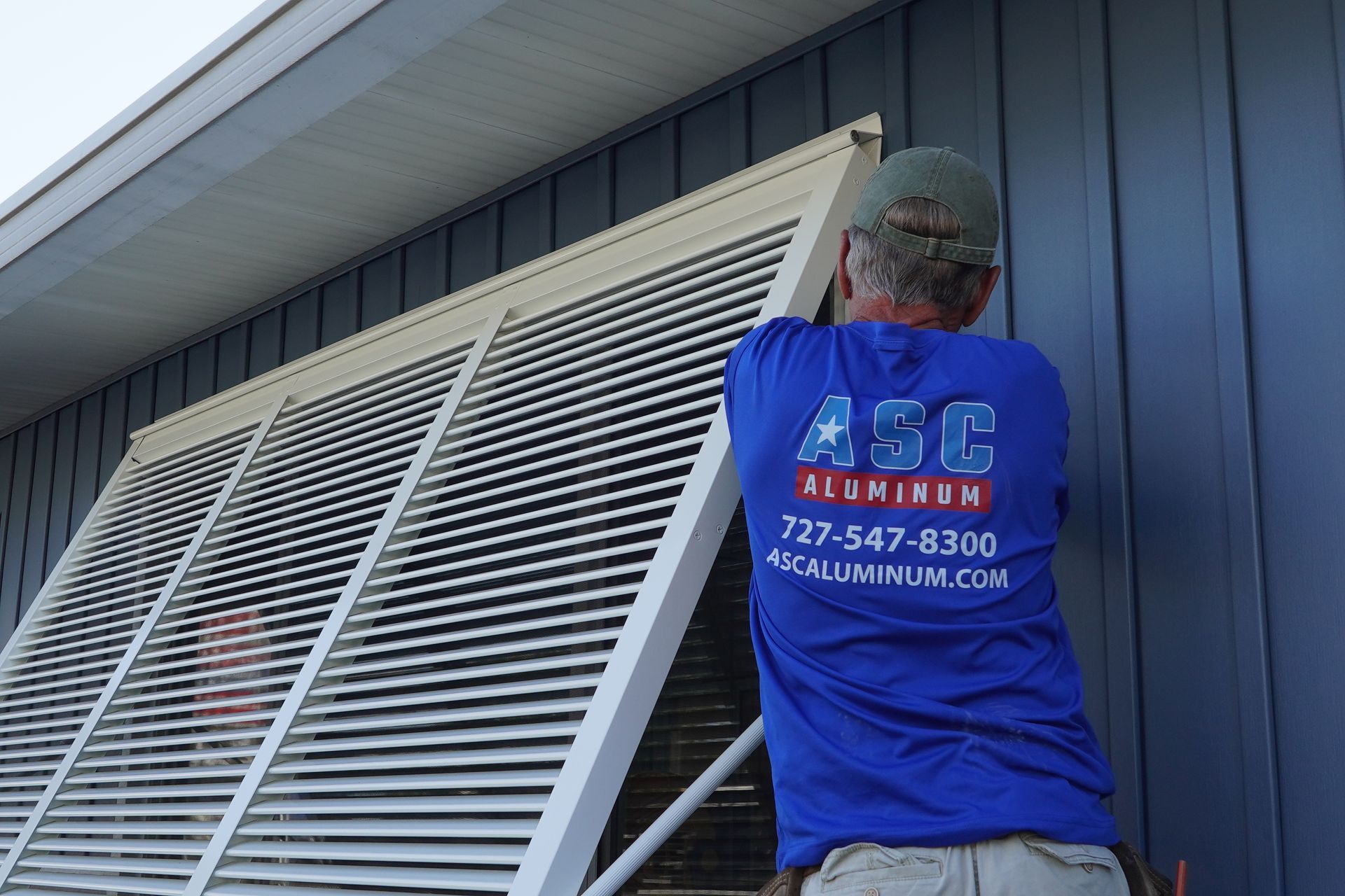 Worker in blue shirt installing a white window shutter on a blue house exterior