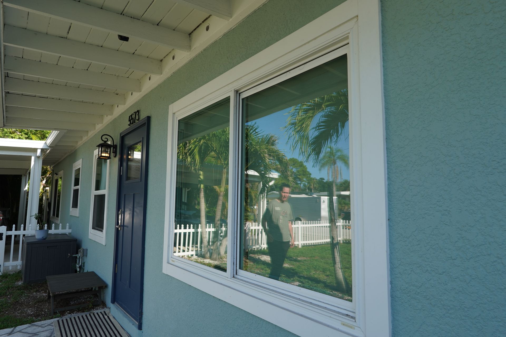 Porch with blue siding, white trim, and large windows reflecting palm trees and a person taking the photo