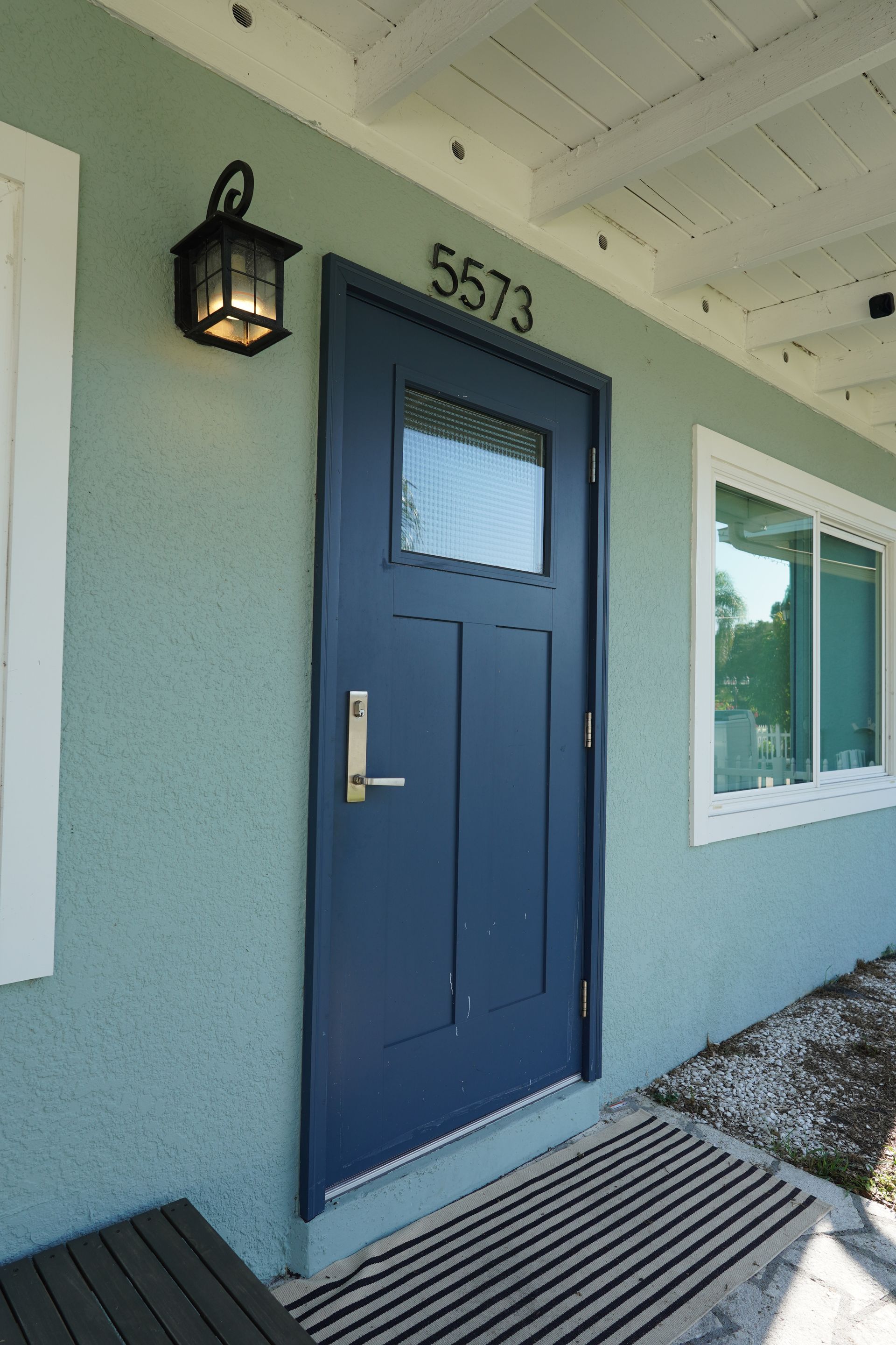Blue front door with numbered entry, wall lantern, and window on a light blue exterior wall