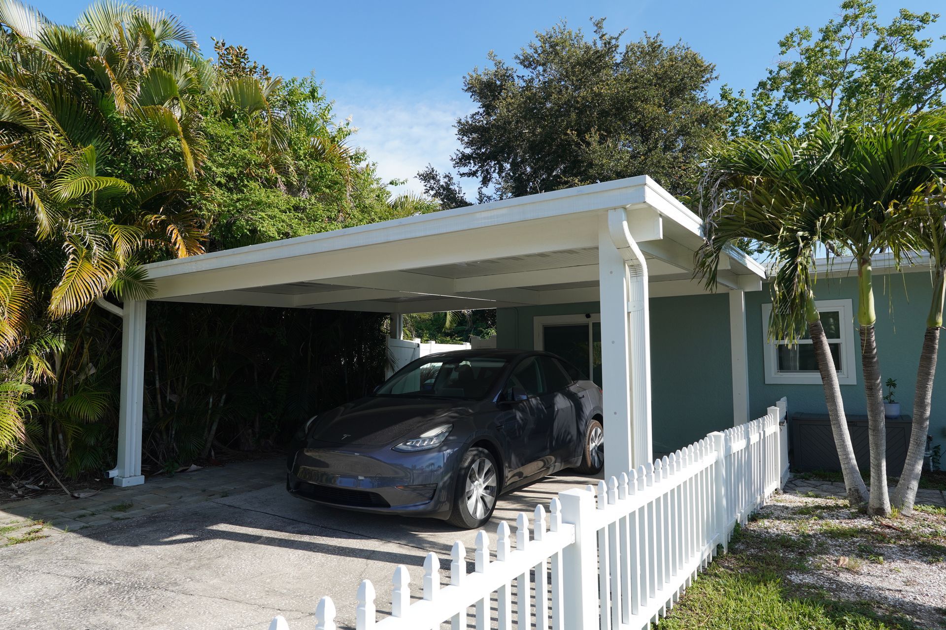 Carport with a black car under a white roof beside a white fence and tropical trees