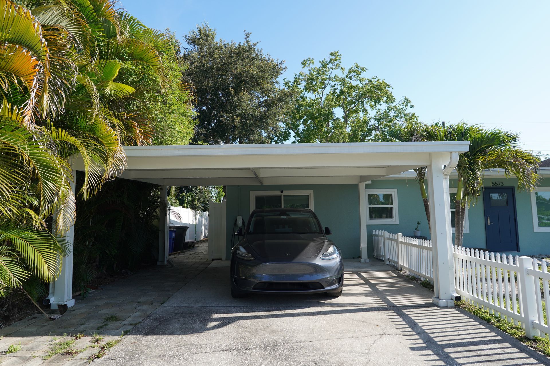 Carport with a parked gray car, white fence, and tropical greenery beside a blue house
