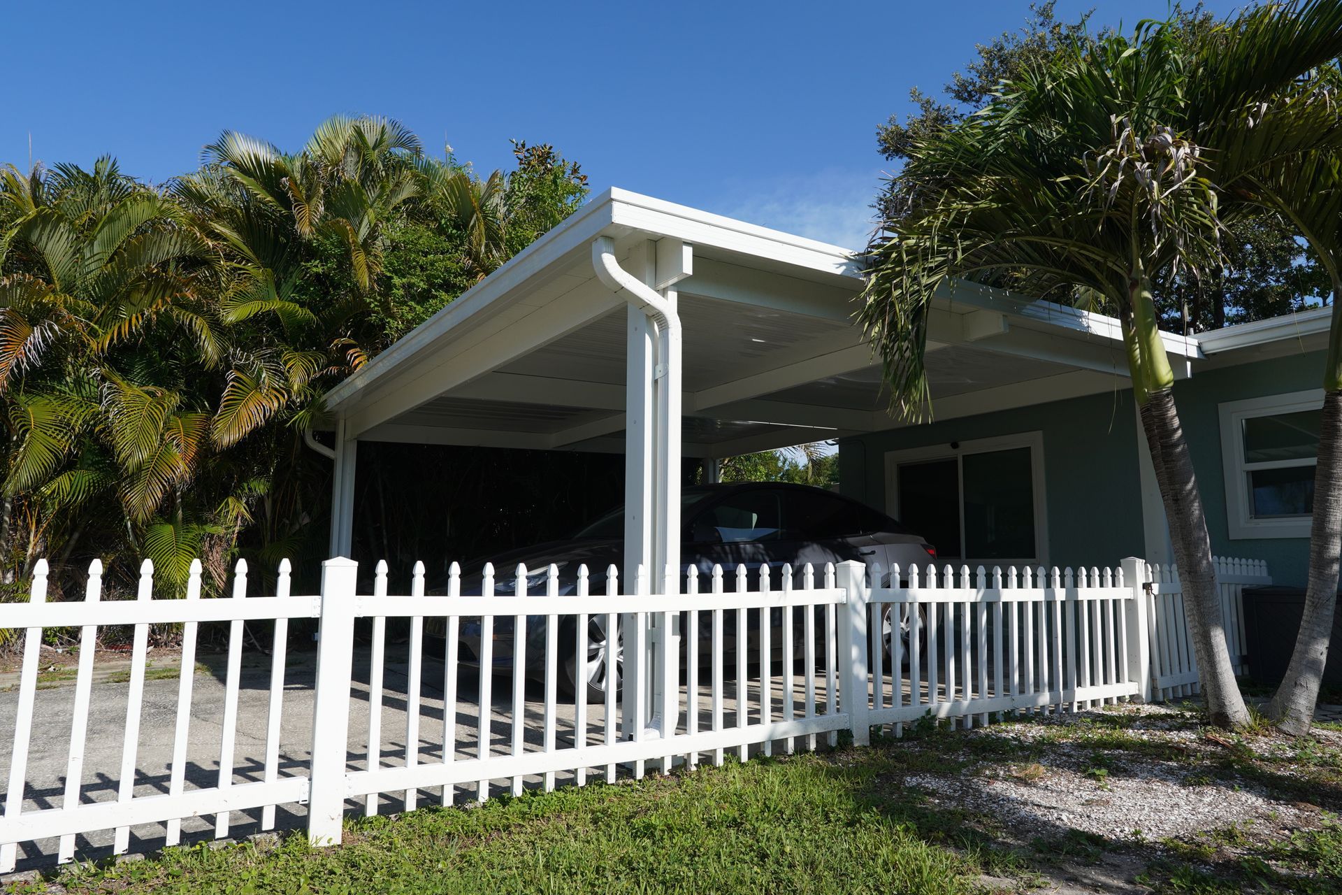 White picket fence in front of a shaded carport with palm trees and a blue house under a sunny sky