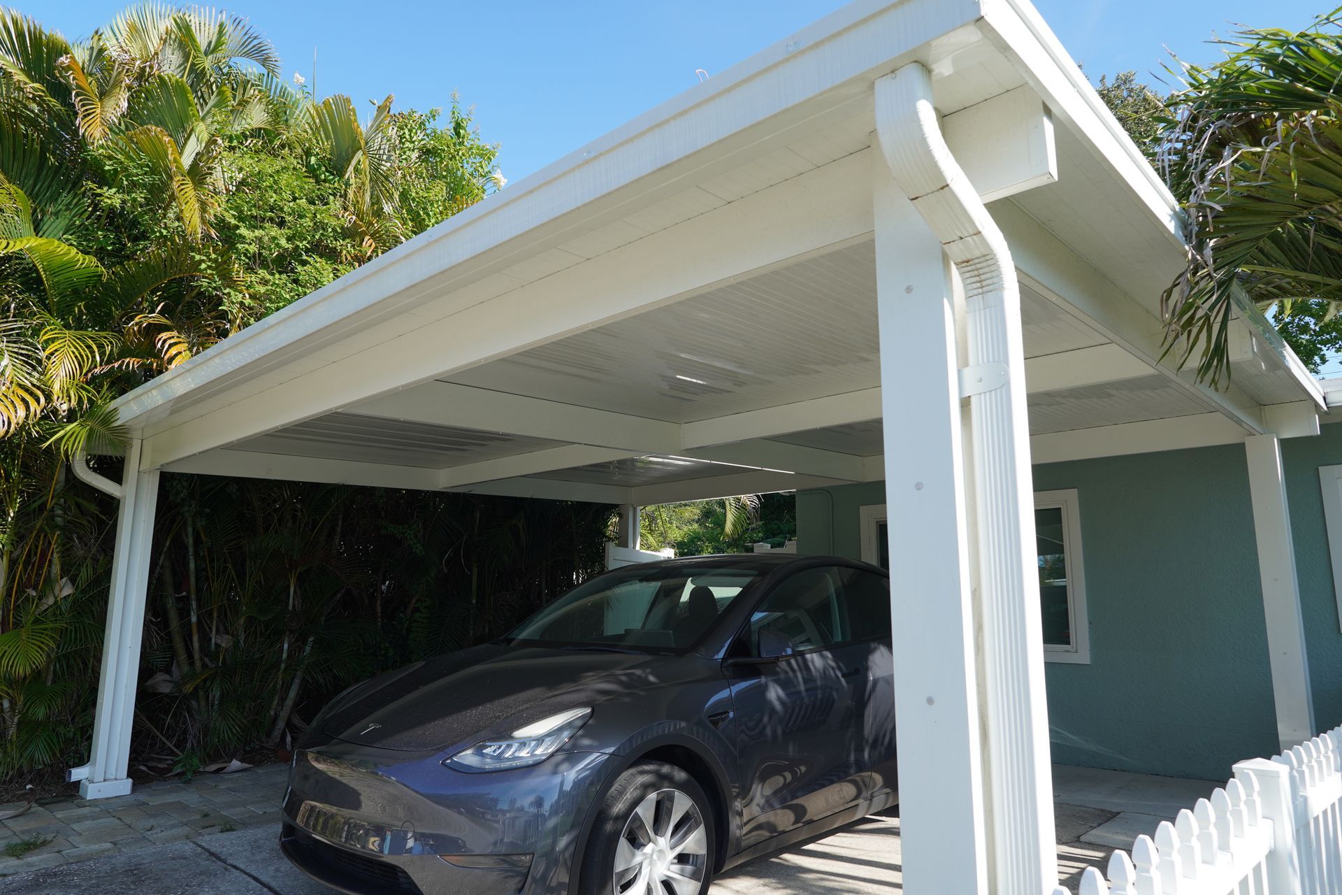 White carport attached to a blue house, with a dark SUV parked underneath and tropical greenery nearby.
