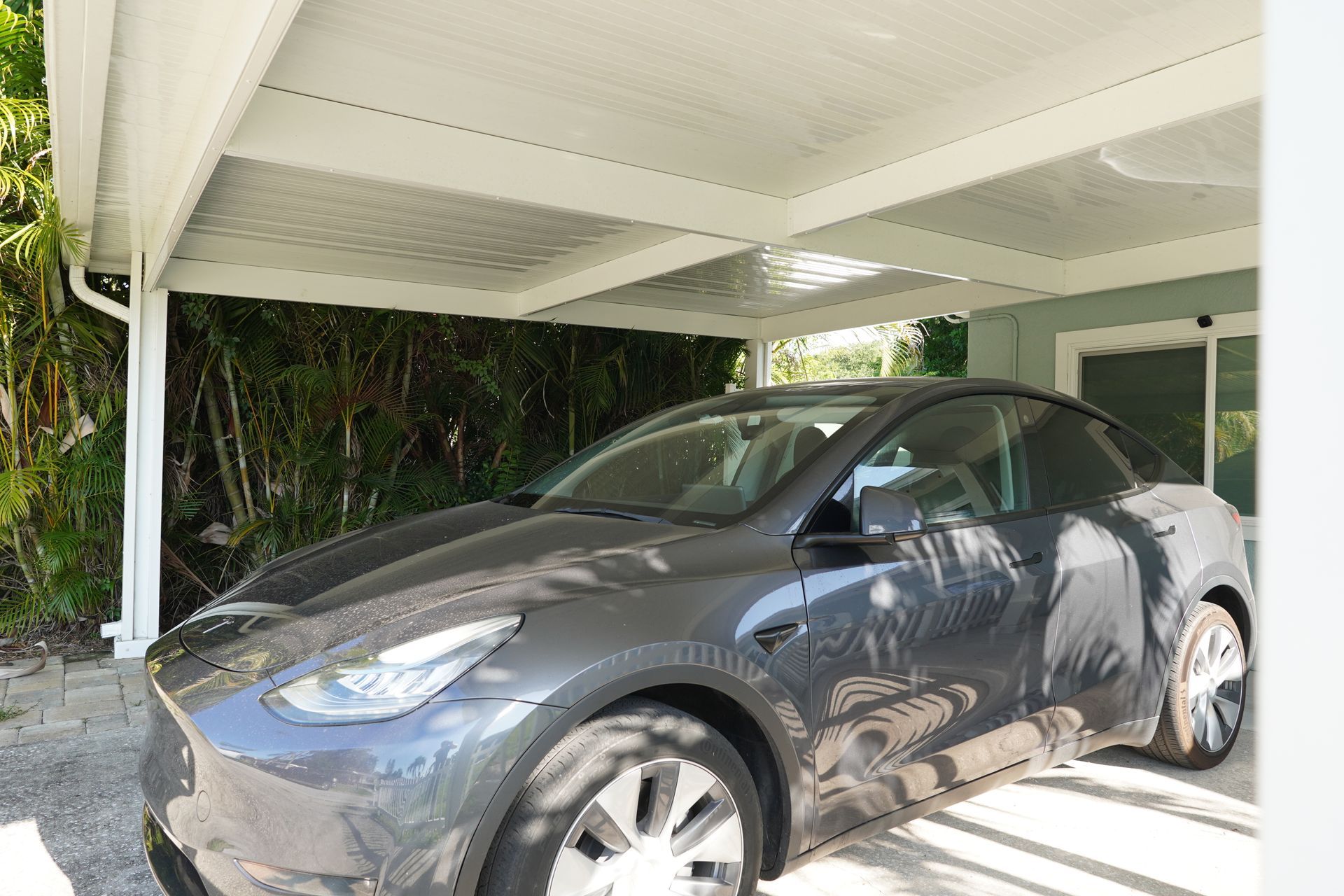 Gray Tesla parked under a white carport with greenery in the background