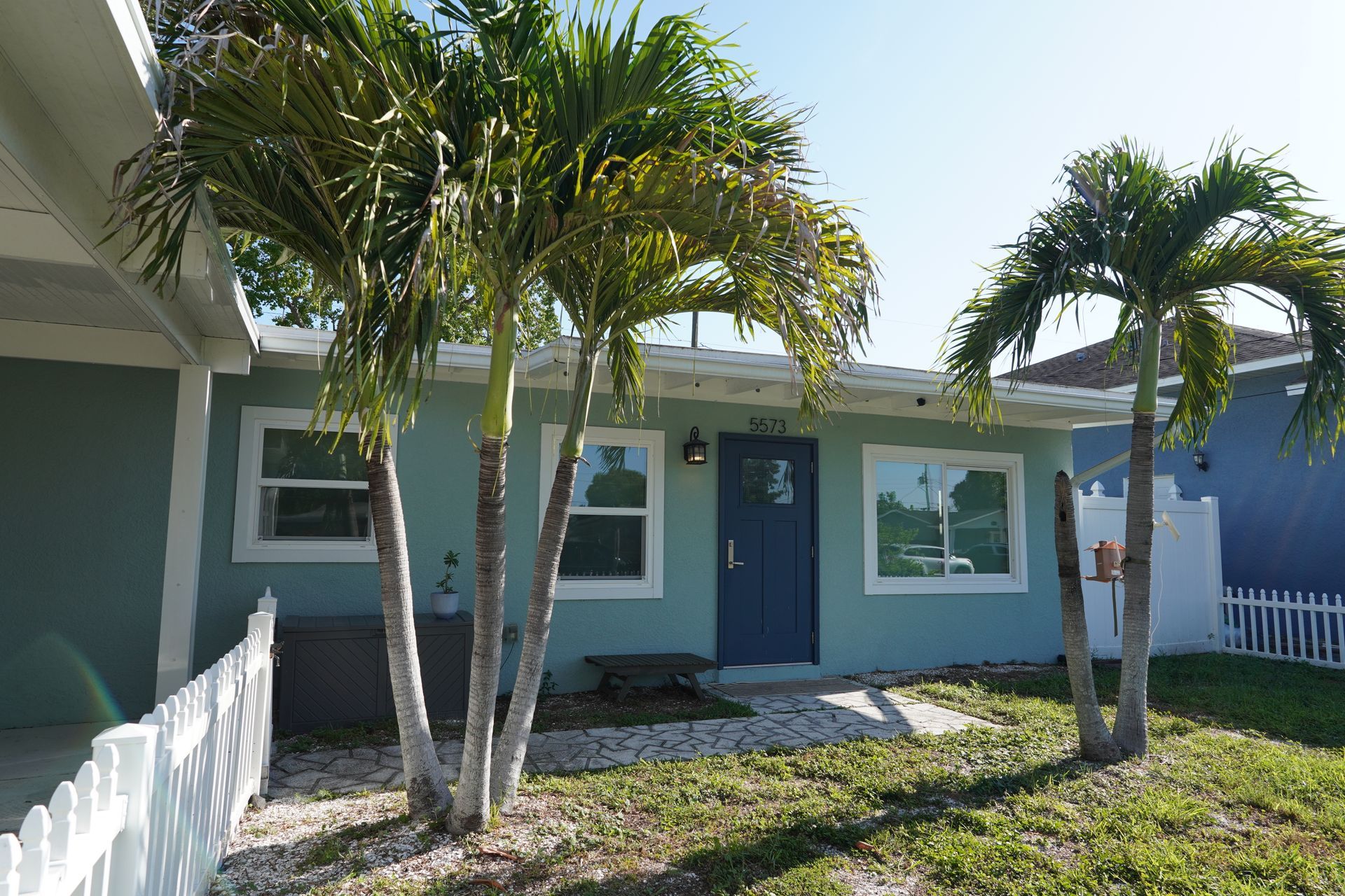 Single-story teal house with white trim, palm trees, and a small front porch in bright sunlight