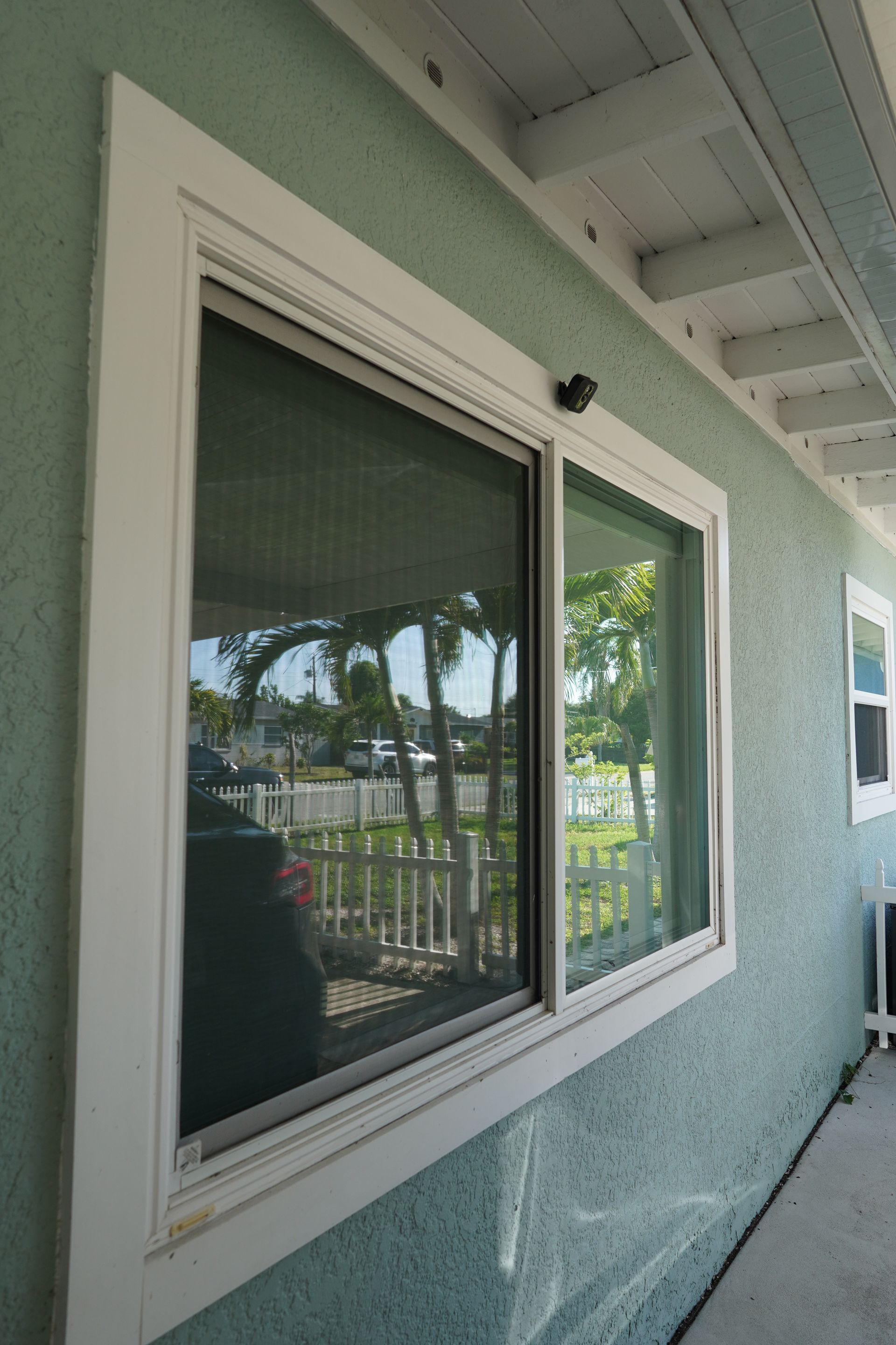 Exterior white-framed window on a mint stucco wall, reflecting palm trees and a railing.