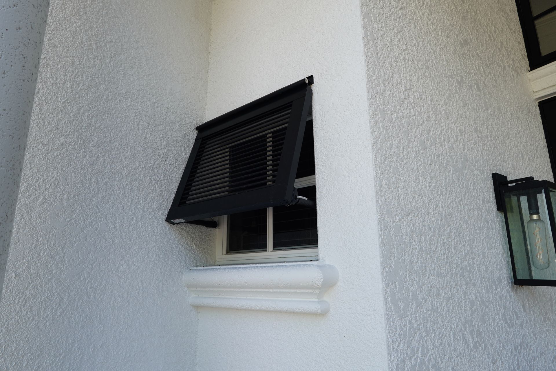 Black awning window partly open on a white exterior wall with a small lantern nearby