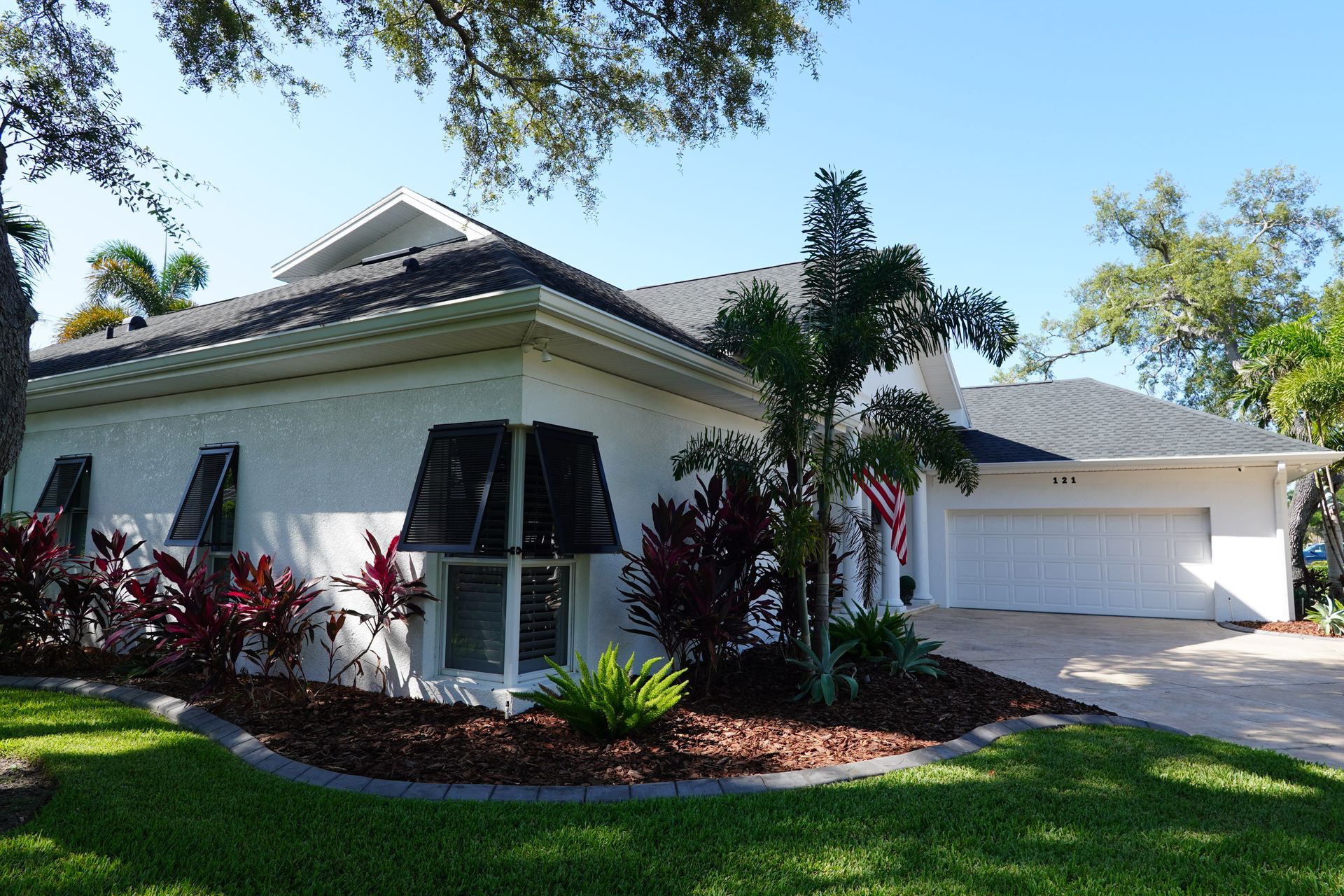 Single-story white house with black shutters, attached garage, and landscaped front yard under sunny trees