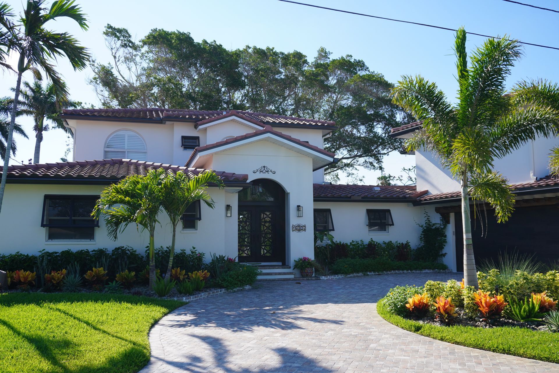 White house with red-tile roof, arched entry, palm trees, and curved driveway in a sunny tropical yard