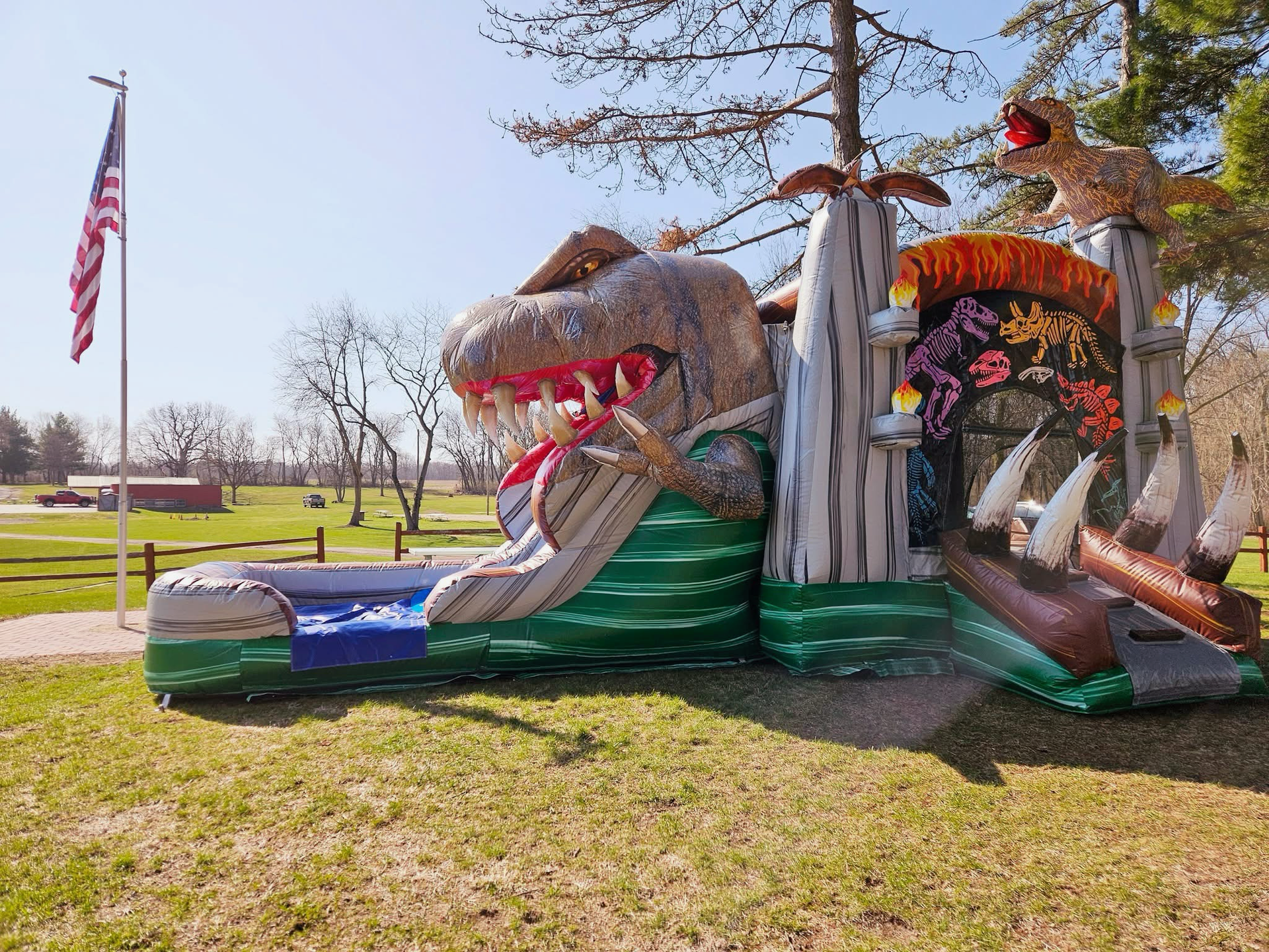 A large, inflatable dinosaur-themed bouncy castle with a slide sits on a grassy field under a sunny blue sky.