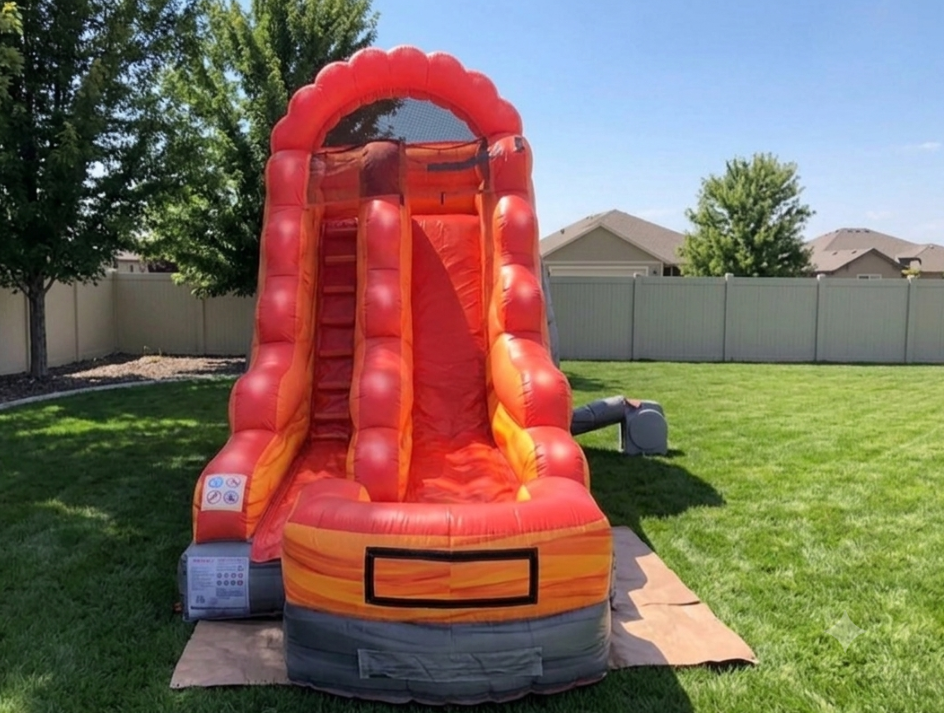Orange inflatable slide in a sunny backyard on green grass, with a tan fence and trees behind it.