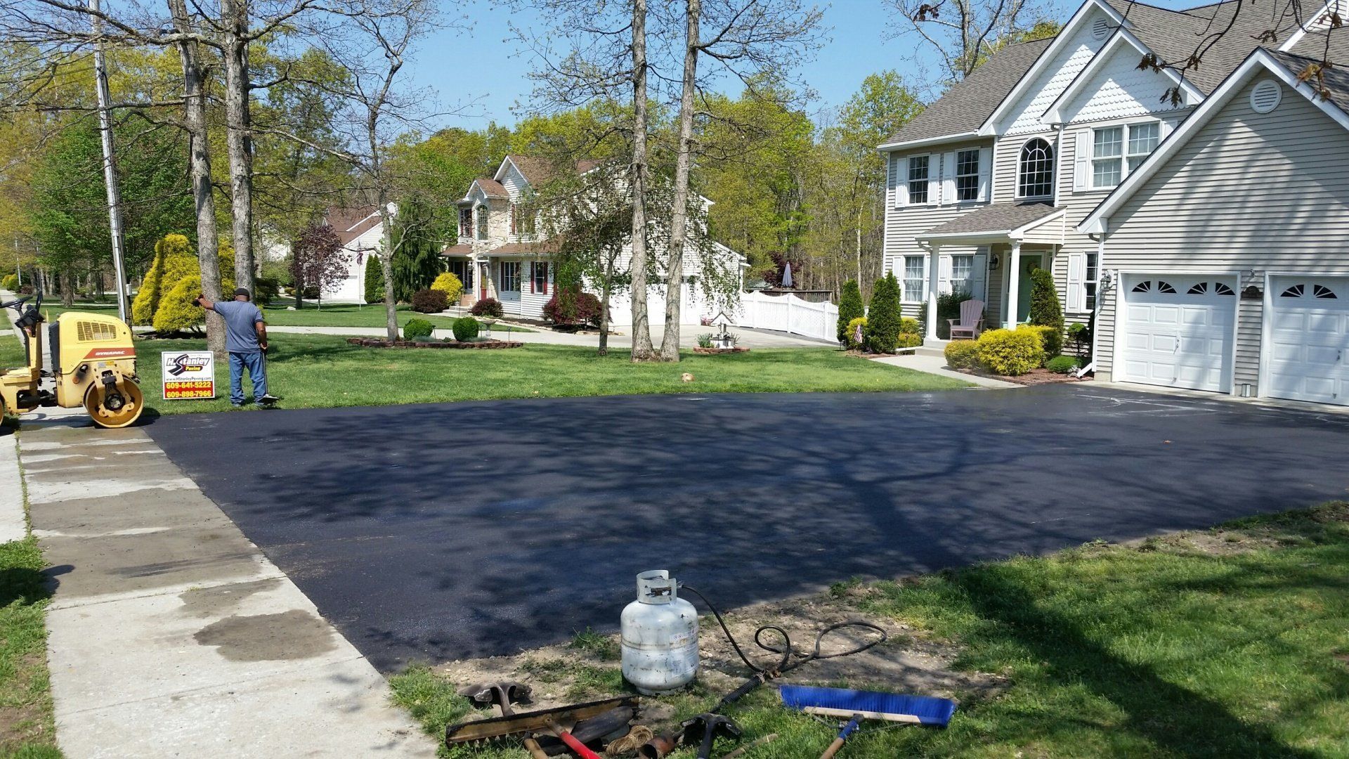 A person stands near a construction roller on a freshly paved black asphalt driveway in front of a suburban house.