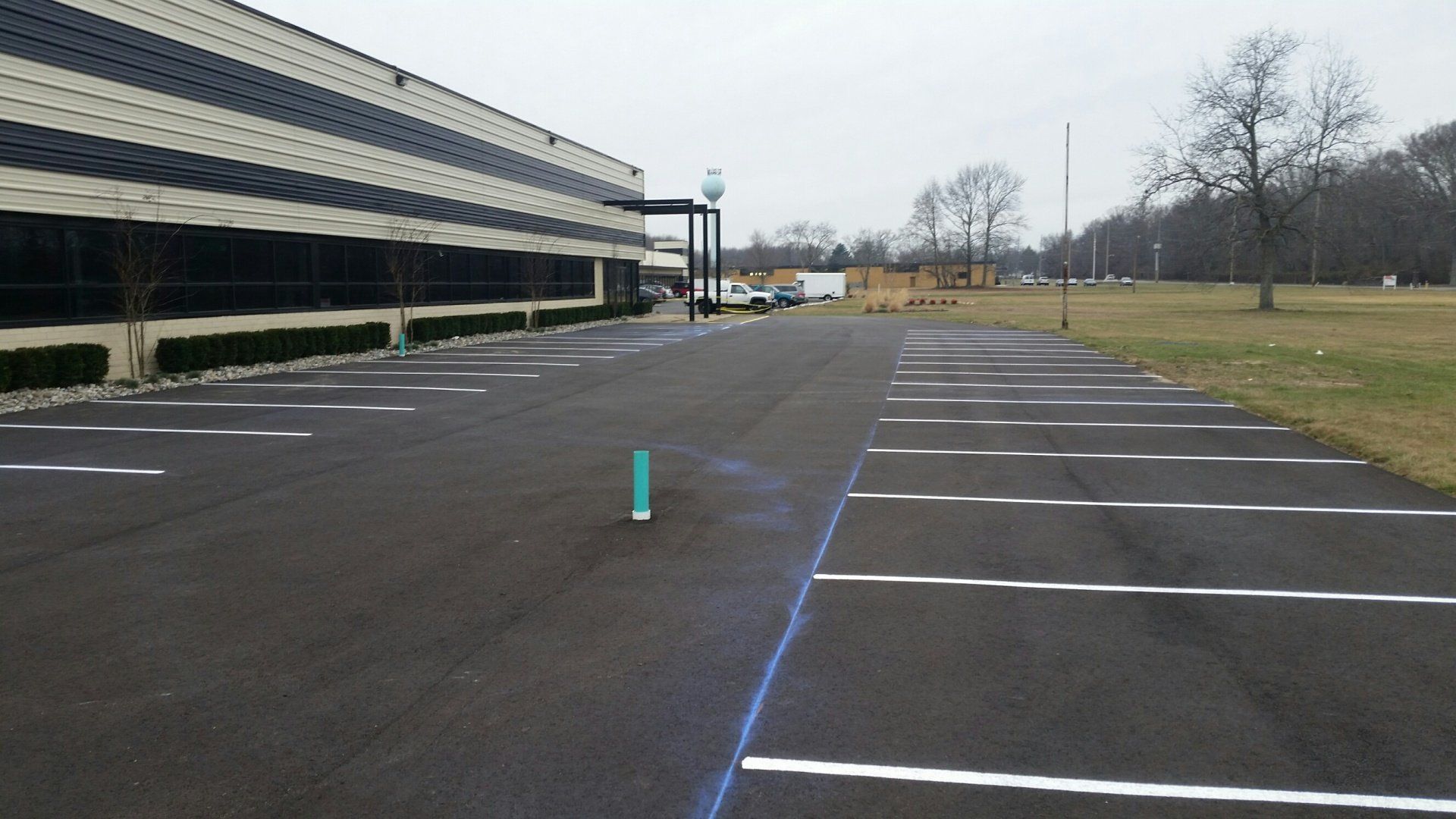 An empty asphalt parking lot with white-painted lines alongside a low building and a grassy area on an overcast day.