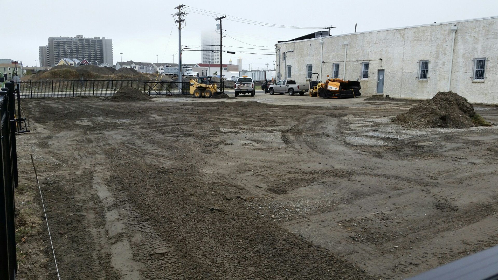 A construction site with piles of dirt, tire tracks, and heavy machinery parked near a long, white industrial building.