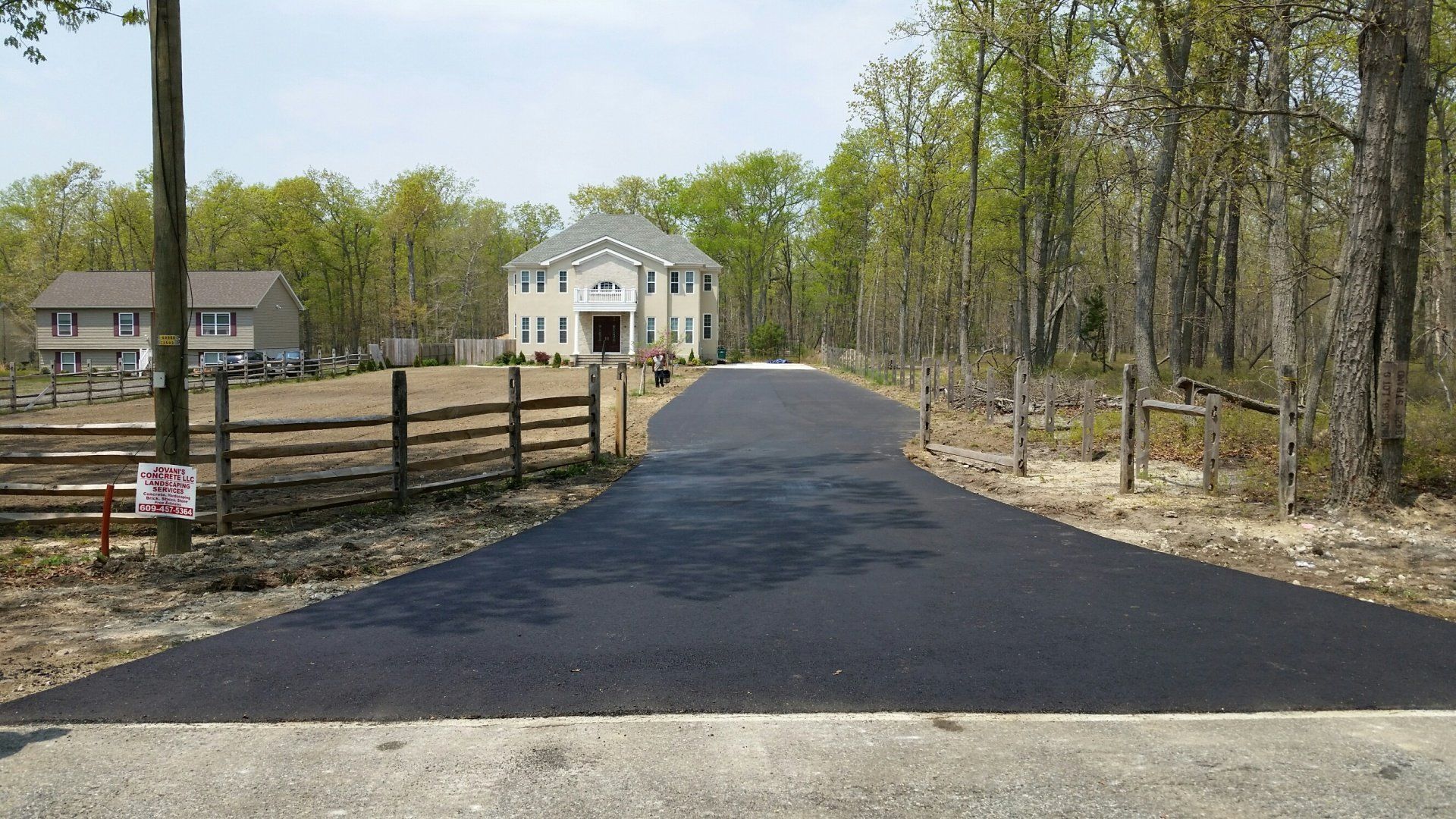 A newly paved black asphalt driveway leads toward a two-story beige house surrounded by woods and a wooden fence.