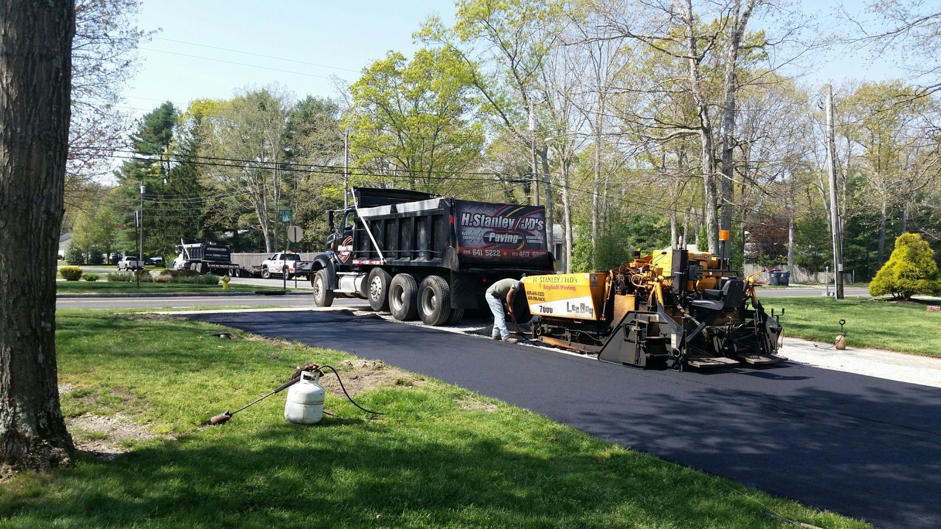 A dump truck and a paver working on a fresh black asphalt driveway in a residential, tree-lined neighborhood.