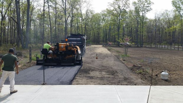 A crew uses a black asphalt paver to lay a new driveway through a wooded residential area.