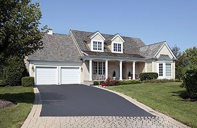 A two-story beige house with a grey roof, white garage doors, and a paved driveway, surrounded by a lawn and trees.