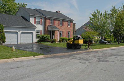 A yellow construction roller sits on the driveway of a two-story brick and siding house on a sunny day.