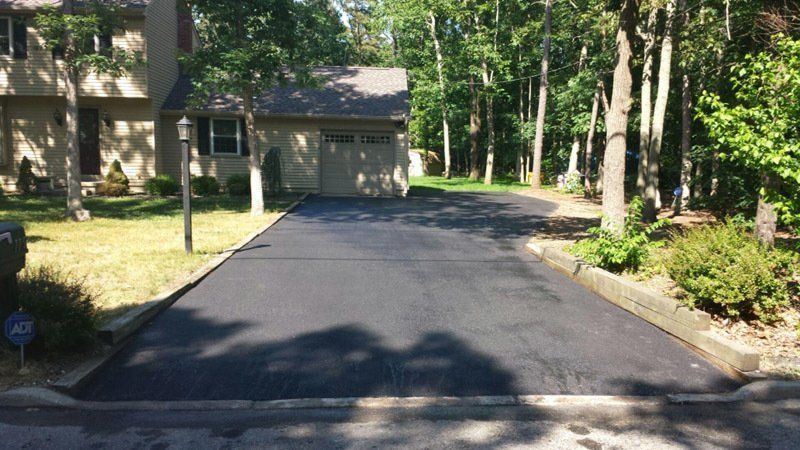 A newly paved dark asphalt driveway leads to a suburban house with a garage, bordered by wooden beams and surrounding trees.