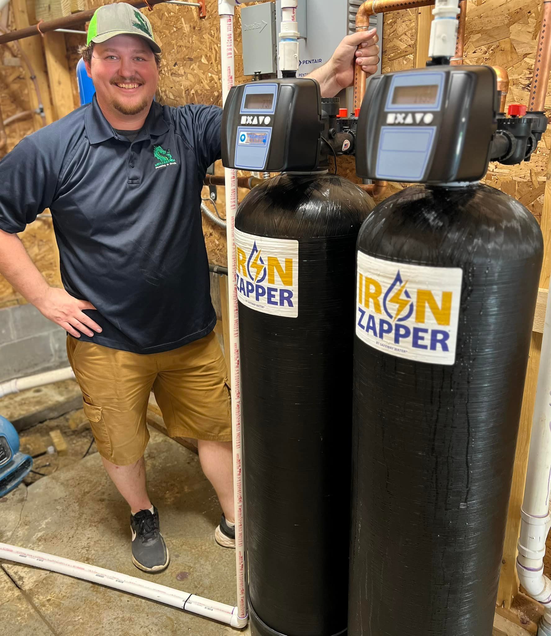 Man stands next to two black water filter tanks, smiling. He wears a polo, shorts, and a green cap, in a utility room.